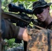 A young male soldier in MARPAT (Marine pattern) camouflage uniform is aiming a scoped rifle in a jungle or forested environment. He is wearing tactical eyewear pushed up on his face and a camouflage patrol cap. The image is taken from a low, close-up angle emphasizing the weapon and the shooter's focused expression.