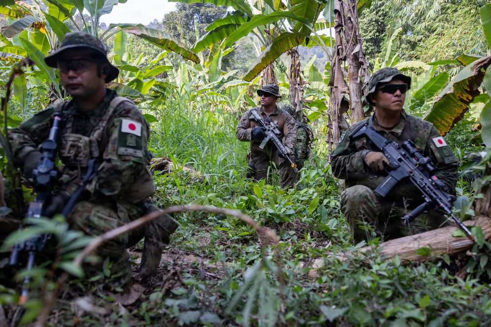 Three armed soldiers are positioned in a dense tropical jungle environment with banana trees and lush vegetation. At least two of the soldiers display Japanese flag patches on their uniforms, identifying them as members of the Japan Ground Self-Defense Force (JGSDF). The soldiers appear to be conducting a patrol or tactical exercise in a jungle terrain.