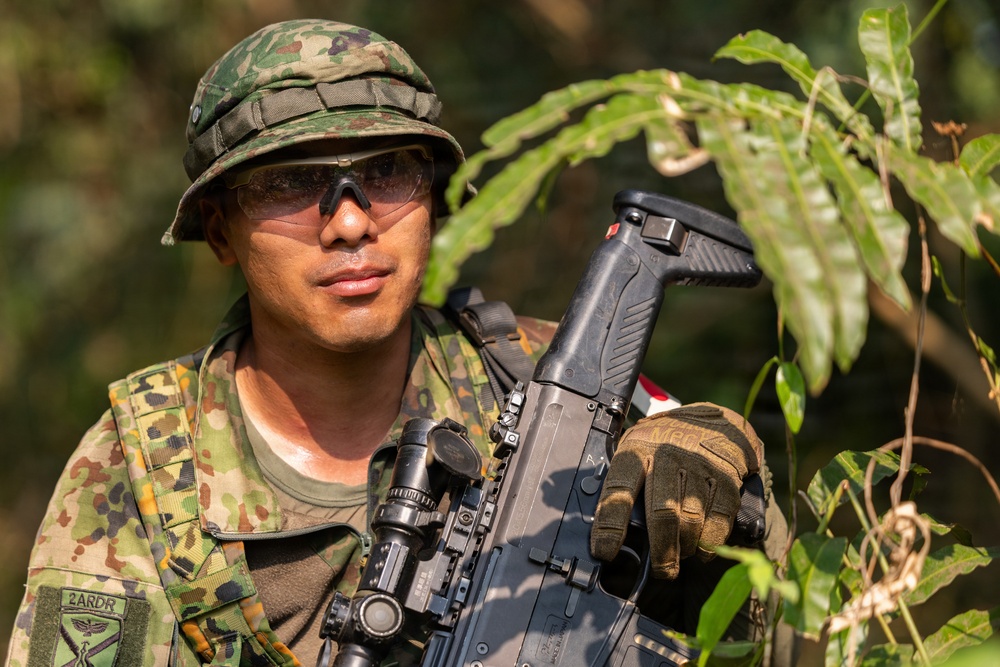 A soldier in full camouflage uniform is photographed in an outdoor jungle or forested environment. He is equipped with tactical gear including a camouflage boonie hat, protective eyewear, tactical gloves, and is holding an assault rifle with optical attachments. A unit patch reading '2ARDR' with an eagle emblem is visible on his left sleeve.