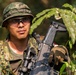 A soldier in full camouflage uniform is photographed in an outdoor jungle or forested environment. He is equipped with tactical gear including a camouflage boonie hat, protective eyewear, tactical gloves, and is holding an assault rifle with optical attachments. A unit patch reading '2ARDR' with an eagle emblem is visible on his left sleeve.