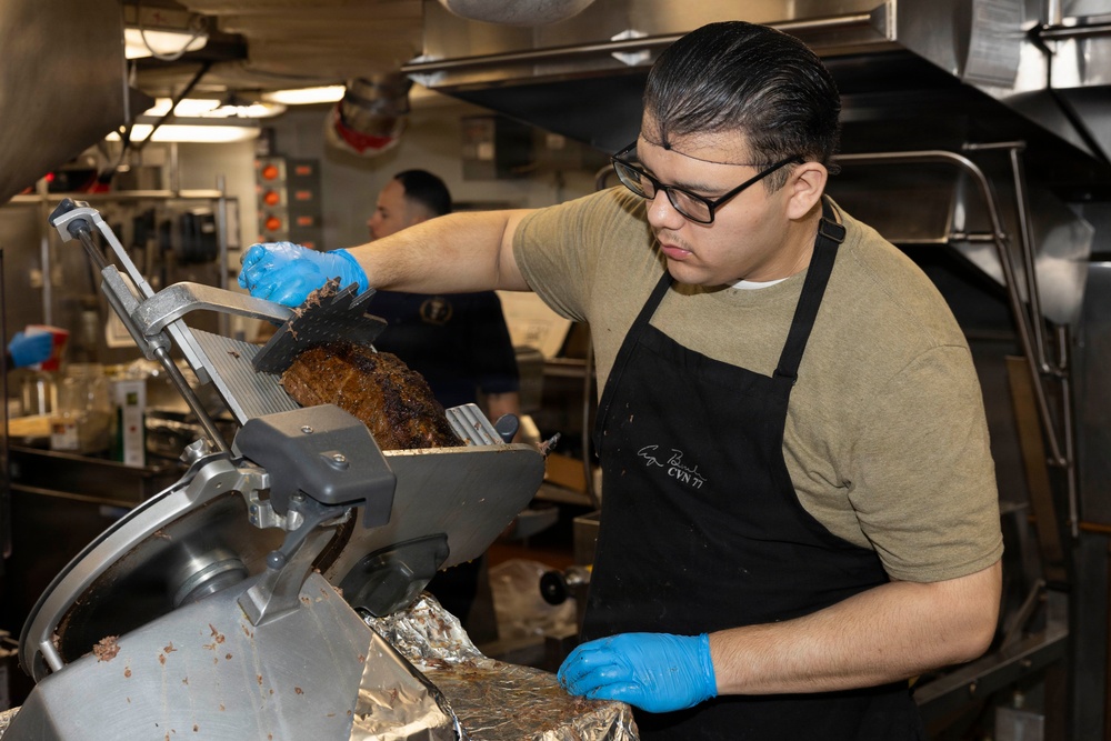 A food service worker wearing a black apron, glasses, and blue nitrile gloves is slicing a large piece of roasted or smoked meat using an industrial meat slicer. The apron appears to have text reading 'CVN 71', suggesting this may be a U.S. Navy ship galley (USS Theodore Roosevelt). A second worker is visible in the background of what appears to be a commercial or military kitchen.