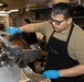A food service worker wearing a black apron, glasses, and blue nitrile gloves is slicing a large piece of roasted or smoked meat using an industrial meat slicer. The apron appears to have text reading 'CVN 71', suggesting this may be a U.S. Navy ship galley (USS Theodore Roosevelt). A second worker is visible in the background of what appears to be a commercial or military kitchen.
