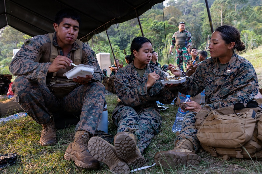 Three military personnel in MARPAT (Marine pattern) camouflage uniforms are seated on grass under a tarp or canopy, eating meals from plates and containers. In the background, additional personnel in different camouflage uniforms (appearing to be a different military branch or nation) are visible standing and socializing. The setting appears to be an outdoor field environment surrounded by tropical vegetation, suggesting a joint military exercise or training event.