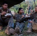 Three military personnel in MARPAT (Marine pattern) camouflage uniforms are seated on grass under a tarp or canopy, eating meals from plates and containers. In the background, additional personnel in different camouflage uniforms (appearing to be a different military branch or nation) are visible standing and socializing. The setting appears to be an outdoor field environment surrounded by tropical vegetation, suggesting a joint military exercise or training event.