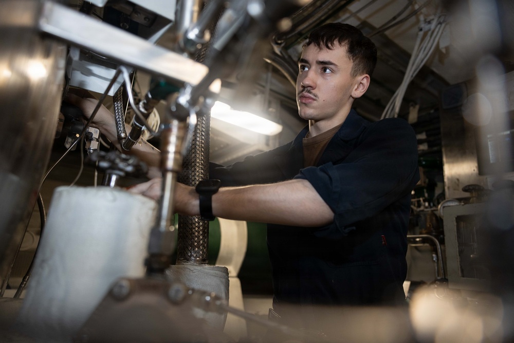 A young male technician or military enlisted personnel is working on complex mechanical or engineering equipment in a confined, industrial space. He is wearing a dark navy blue uniform or coveralls with a brown undershirt and a black wristband, suggesting a military or naval maintenance role. The environment appears to be an engine room or mechanical compartment, likely aboard a naval vessel.