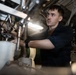 A young male technician or military enlisted personnel is working on complex mechanical or engineering equipment in a confined, industrial space. He is wearing a dark navy blue uniform or coveralls with a brown undershirt and a black wristband, suggesting a military or naval maintenance role. The environment appears to be an engine room or mechanical compartment, likely aboard a naval vessel.