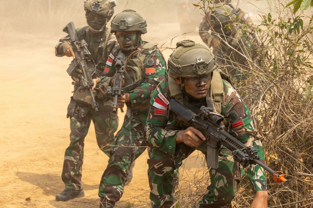 The image shows a group of Indonesian military soldiers conducting what appears to be a tactical field exercise or training operation. The soldiers are wearing camouflage uniforms with visible 'INDONESIA' patches and red-and-white flag insignia, moving through a dusty, semi-arid terrain with sparse vegetation. They are armed and moving in a tactical formation, suggesting a coordinated military drill.