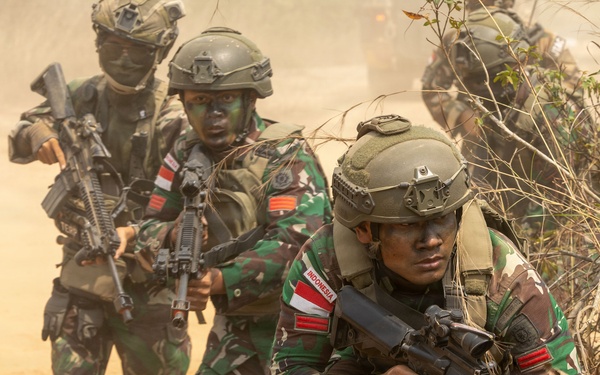 The image shows a group of Indonesian military soldiers conducting what appears to be a tactical field exercise or training operation. The soldiers are wearing camouflage uniforms with visible 'INDONESIA' patches and red-and-white flag insignia, moving through a dusty, semi-arid terrain with sparse vegetation. They are armed and moving in a tactical formation, suggesting a coordinated military drill.