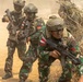The image shows a group of Indonesian military soldiers conducting what appears to be a tactical field exercise or training operation. The soldiers are wearing camouflage uniforms with visible 'INDONESIA' patches and red-and-white flag insignia, moving through a dusty, semi-arid terrain with sparse vegetation. They are armed and moving in a tactical formation, suggesting a coordinated military drill.