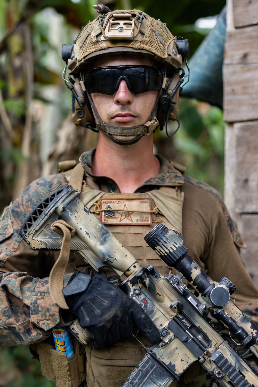 A U.S. Marine in full combat gear is photographed in what appears to be a tropical environment. The Marine is equipped with advanced tactical equipment including a FAST helmet with accessories, plate carrier, and an assault rifle. Name tape and unit patches are visible on the plate carrier.