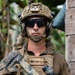 A U.S. Marine in full combat gear is photographed in what appears to be a tropical environment. The Marine is equipped with advanced tactical equipment including a FAST helmet with accessories, plate carrier, and an assault rifle. Name tape and unit patches are visible on the plate carrier.