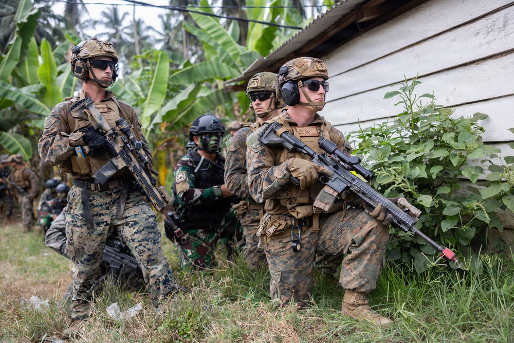 A group of armed military personnel in tactical gear are positioned near a wooden structure in a tropical environment with banana trees and dense vegetation. The group appears to include soldiers in different camouflage patterns, suggesting a multinational or joint military exercise. Personnel are holding rifles and appear to be in a tactical formation or patrol posture.