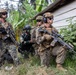 A group of armed military personnel in tactical gear are positioned near a wooden structure in a tropical environment with banana trees and dense vegetation. The group appears to include soldiers in different camouflage patterns, suggesting a multinational or joint military exercise. Personnel are holding rifles and appear to be in a tactical formation or patrol posture.