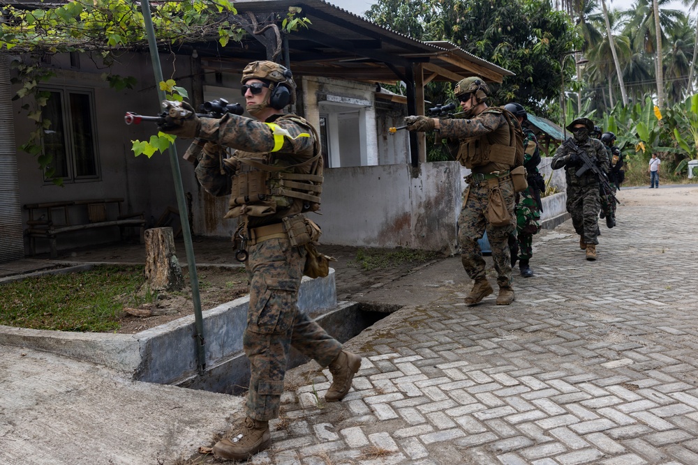 A group of military personnel in full combat gear are conducting what appears to be a training exercise in an urban environment. The soldiers, wearing MARPAT (Marine pattern) camouflage and tactical vests, are moving in a tactical formation along a brick-paved road past civilian-style buildings. The presence of yellow safety armbands and blank-firing adapters on weapons strongly indicates this is a military training exercise rather than a live combat operation.