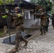 A group of military personnel in full combat gear are conducting what appears to be a training exercise in an urban environment. The soldiers, wearing MARPAT (Marine pattern) camouflage and tactical vests, are moving in a tactical formation along a brick-paved road past civilian-style buildings. The presence of yellow safety armbands and blank-firing adapters on weapons strongly indicates this is a military training exercise rather than a live combat operation.