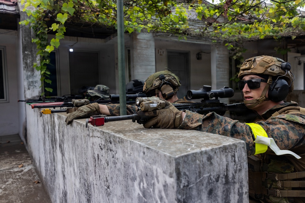 Three military personnel in camouflage uniforms are positioned behind a concrete wall, aiming rifles in a coordinated firing line. The soldiers are equipped with tactical gear including helmets, hearing protection, and optical scopes. The setting appears to be an urban training environment with a courtyard and building structures visible in the background.