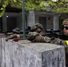 Three military personnel in camouflage uniforms are positioned behind a concrete wall, aiming rifles in a coordinated firing line. The soldiers are equipped with tactical gear including helmets, hearing protection, and optical scopes. The setting appears to be an urban training environment with a courtyard and building structures visible in the background.