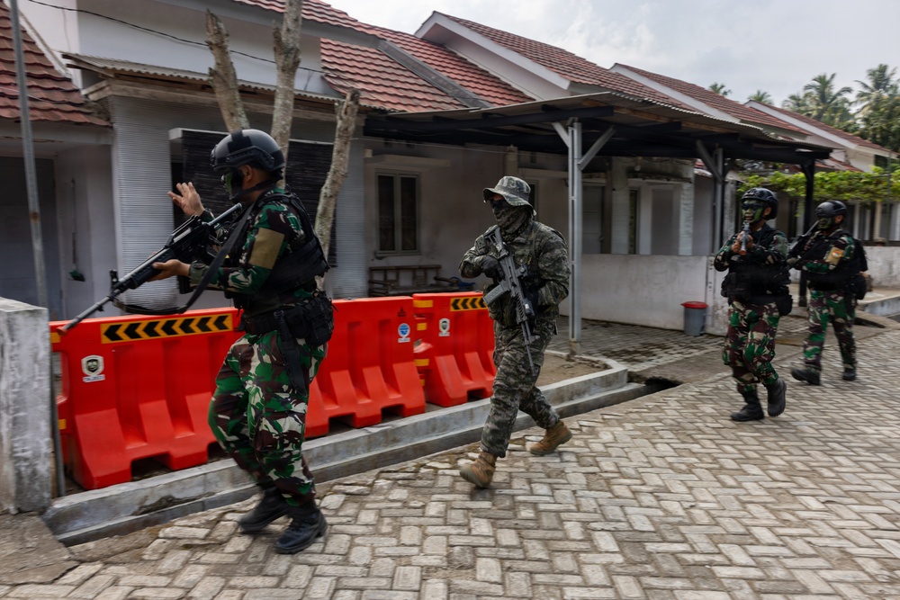 Four armed military or security personnel are moving tactically along a paved pathway between low-rise buildings with red-tiled roofs. Three individuals wear Indonesian military (TNI) camouflage uniforms with helmets and tactical vests, while one wears a different camouflage pattern with a boonie hat and face covering. The scene appears to depict a military exercise or security operation, with orange traffic barriers visible in the background.