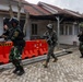 Four armed military or security personnel are moving tactically along a paved pathway between low-rise buildings with red-tiled roofs. Three individuals wear Indonesian military (TNI) camouflage uniforms with helmets and tactical vests, while one wears a different camouflage pattern with a boonie hat and face covering. The scene appears to depict a military exercise or security operation, with orange traffic barriers visible in the background.