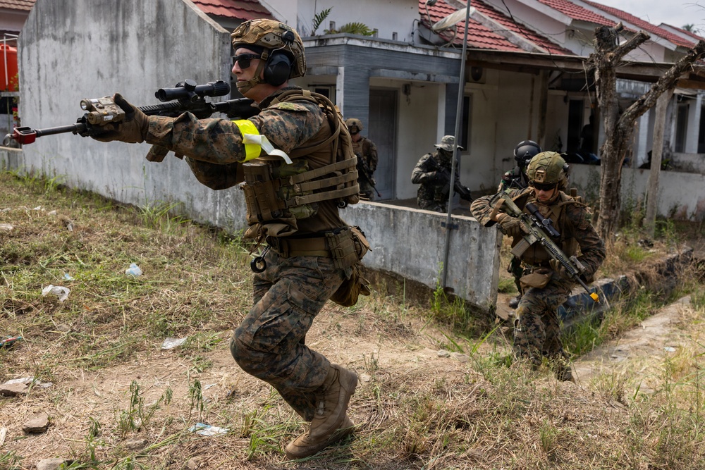 The image shows military personnel in MARPAT (Marine pattern) camouflage uniforms conducting what appears to be a training exercise in an urban environment. The lead soldier is advancing with a rifle raised in a firing position, while teammates follow in tactical formation near a low concrete wall and residential buildings. Yellow armbands and exercise markings suggest this is a simulated combat drill rather than an actual combat operation.