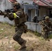 The image shows military personnel in MARPAT (Marine pattern) camouflage uniforms conducting what appears to be a training exercise in an urban environment. The lead soldier is advancing with a rifle raised in a firing position, while teammates follow in tactical formation near a low concrete wall and residential buildings. Yellow armbands and exercise markings suggest this is a simulated combat drill rather than an actual combat operation.