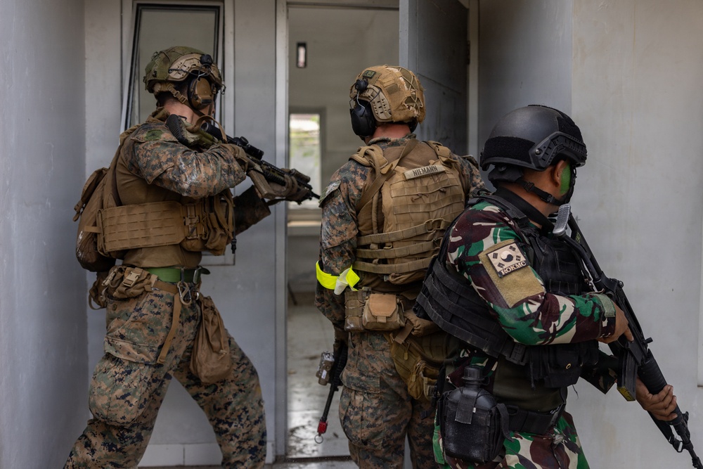 Three armed military personnel are conducting a tactical building entry exercise, stacked near a doorway in a clearing or breach formation. Two soldiers wear US Marine Corps MARPAT digital camouflage uniforms with tan plate carriers, while a third wears a tropical camouflage pattern with a South Korean flag patch visible on the shoulder. The scene appears to be a joint military training exercise involving US Marines and a Korean or allied nation's forces.