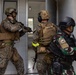 Three armed military personnel are conducting a tactical building entry exercise, stacked near a doorway in a clearing or breach formation. Two soldiers wear US Marine Corps MARPAT digital camouflage uniforms with tan plate carriers, while a third wears a tropical camouflage pattern with a South Korean flag patch visible on the shoulder. The scene appears to be a joint military training exercise involving US Marines and a Korean or allied nation's forces.