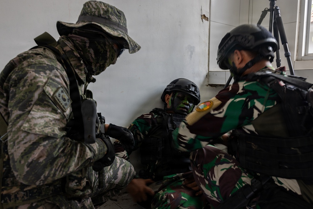Three military personnel are engaged in what appears to be a combat casualty care or tactical medical training exercise inside a building. One soldier wearing green/brown camouflage uniform is seated against a wall in a simulated casualty role, while two others attend to them. The soldiers wear face paint and tactical gear consistent with a joint military training exercise.