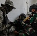Three military personnel are engaged in what appears to be a combat casualty care or tactical medical training exercise inside a building. One soldier wearing green/brown camouflage uniform is seated against a wall in a simulated casualty role, while two others attend to them. The soldiers wear face paint and tactical gear consistent with a joint military training exercise.