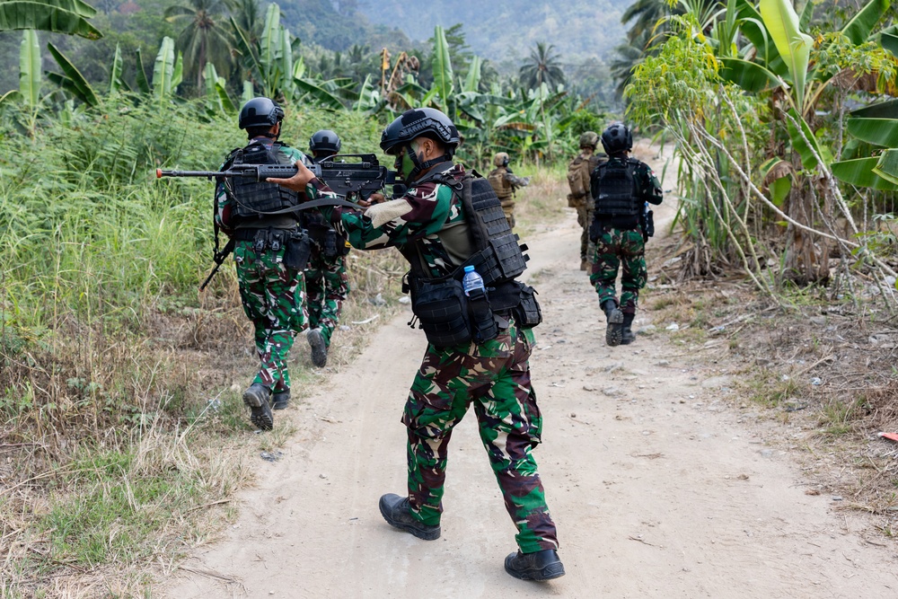 A group of armed military or special operations personnel are conducting a patrol along a dirt trail through a tropical jungle environment. The soldiers are wearing camouflage uniforms with tactical vests and helmets, carrying assault rifles in a ready position. The vegetation and uniform patterns suggest a Southeast Asian operational context.