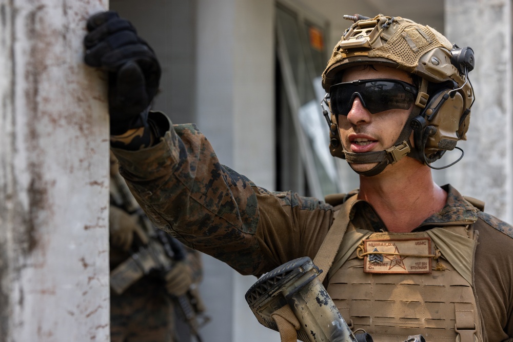 A U.S. Marine in full combat gear is positioned against a wall, appearing to be conducting urban warfare training or an operational exercise. The Marine is wearing MARPAT camouflage, a tan plate carrier with visible name tapes reading 'MURRAY' and 'USMC', and a tactical helmet with communications equipment. The setting appears to be an urban or built environment.