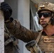 A U.S. Marine in full combat gear is positioned against a wall, appearing to be conducting urban warfare training or an operational exercise. The Marine is wearing MARPAT camouflage, a tan plate carrier with visible name tapes reading 'MURRAY' and 'USMC', and a tactical helmet with communications equipment. The setting appears to be an urban or built environment.