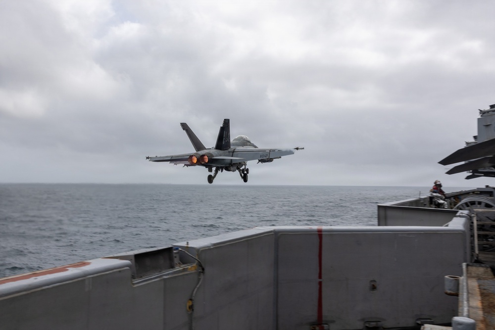 A U.S. Navy carrier-based jet aircraft, marked with the number '102', is captured mid-launch or approach over the flight deck of an aircraft carrier at sea. The aircraft's afterburners are visibly ignited, and its landing gear is deployed. The scene takes place under overcast skies over open ocean.
