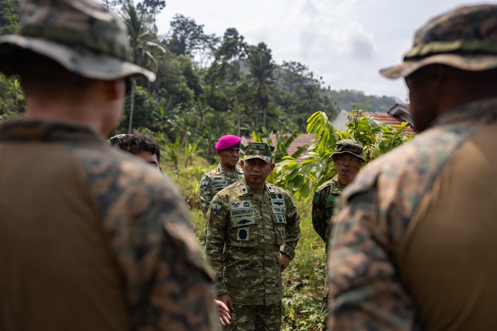 A group of military personnel from multiple nations are gathered in an outdoor briefing or discussion in a tropical, vegetated environment. The central figure wears a name tape reading 'ANGGOROJATI' with 'TNI AL' (Indonesian Navy) insignia, while others in the foreground wear US Marine Corps MARPAT camouflage uniforms. This appears to be a joint multinational military exercise or coordination meeting.