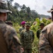 A group of military personnel from multiple nations are gathered in an outdoor briefing or discussion in a tropical, vegetated environment. The central figure wears a name tape reading 'ANGGOROJATI' with 'TNI AL' (Indonesian Navy) insignia, while others in the foreground wear US Marine Corps MARPAT camouflage uniforms. This appears to be a joint multinational military exercise or coordination meeting.