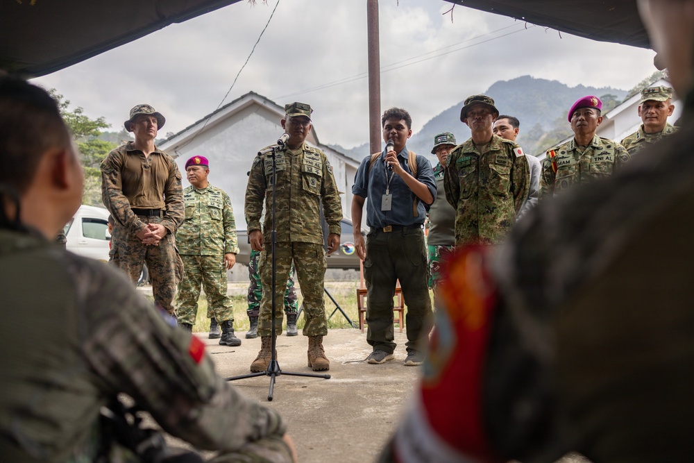 A multinational military briefing or ceremony is taking place outdoors under a canopy structure, with personnel from multiple nations gathered together. A central figure in camouflage uniform stands at a microphone stand while a civilian interpreter holds a handheld microphone nearby. The gathering includes personnel wearing distinctly different uniforms, suggesting a joint military exercise or cooperative event.