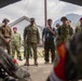 A multinational military briefing or ceremony is taking place outdoors under a canopy structure, with personnel from multiple nations gathered together. A central figure in camouflage uniform stands at a microphone stand while a civilian interpreter holds a handheld microphone nearby. The gathering includes personnel wearing distinctly different uniforms, suggesting a joint military exercise or cooperative event.