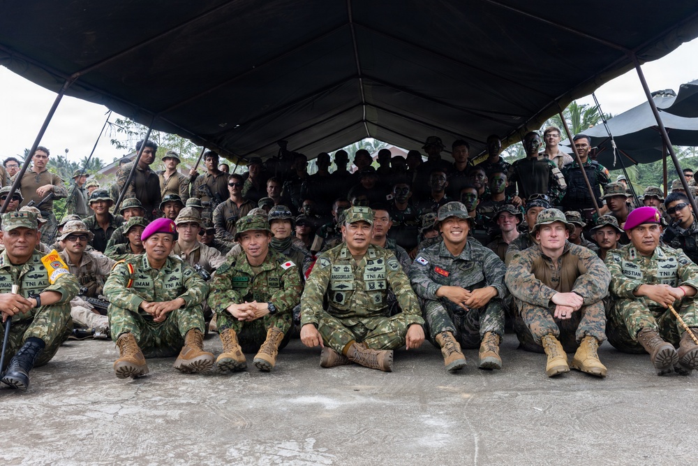 A large multinational military group photo taken under a dark canopy tent, likely during a joint military exercise or training event. Personnel from multiple nations are visible, including identifiable patches and flags from Indonesia (TNI AL), Japan, South Korea, and the United States. Soldiers are arranged in rows, with front-row members seated on the ground and others standing behind them.