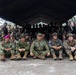 A large multinational military group photo taken under a dark canopy tent, likely during a joint military exercise or training event. Personnel from multiple nations are visible, including identifiable patches and flags from Indonesia (TNI AL), Japan, South Korea, and the United States. Soldiers are arranged in rows, with front-row members seated on the ground and others standing behind them.