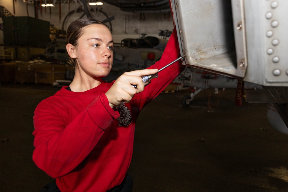 A young person wearing a red sweatshirt is using a screwdriver to perform maintenance on an aircraft panel in what appears to be a military hangar bay. The individual is focused on working on a metal access panel on the underside or side of an aircraft. The setting suggests this is aboard a naval vessel or military air facility.