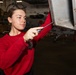 A young person wearing a red sweatshirt is using a screwdriver to perform maintenance on an aircraft panel in what appears to be a military hangar bay. The individual is focused on working on a metal access panel on the underside or side of an aircraft. The setting suggests this is aboard a naval vessel or military air facility.