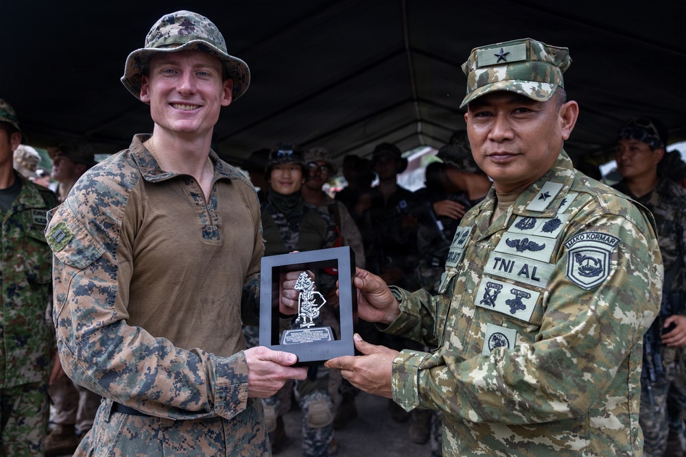 Two military personnel from different nations are jointly holding a commemorative plaque or award featuring a decorative figurine. The officer on the right wears a uniform marked 'TNI AL' (Indonesian Navy - Tentara Nasional Indonesia Angkatan Laut) with a one-star rank insignia, while the officer on the left wears US Marine Corps MARPAT camouflage. This appears to be a ceremonial exchange or award presentation during a joint military exercise.