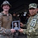 Two military personnel from different nations are jointly holding a commemorative plaque or award featuring a decorative figurine. The officer on the right wears a uniform marked 'TNI AL' (Indonesian Navy - Tentara Nasional Indonesia Angkatan Laut) with a one-star rank insignia, while the officer on the left wears US Marine Corps MARPAT camouflage. This appears to be a ceremonial exchange or award presentation during a joint military exercise.