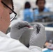 A laboratory technician wearing a white lab coat, black-framed glasses, and gray nitrile gloves is closely examining a small glass slide or specimen containing a red substance, likely blood. The setting appears to be a clinical or research laboratory environment. At least two additional personnel in blue scrubs or lab attire are visible in the blurred background.