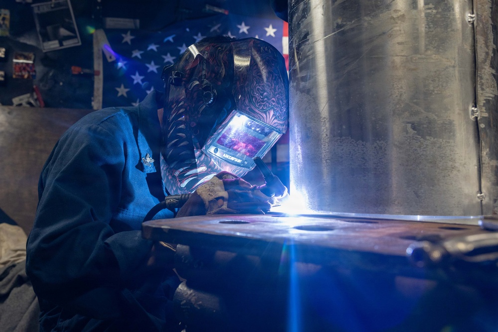 A welder is actively welding at the base of a large cylindrical metal structure, producing a bright arc flash. The worker is wearing a decorative auto-darkening welding helmet branded 'ARCCAPTAIN' and a blue welding jacket. An American flag is visible in the background, suggesting a workshop or fabrication shop setting.