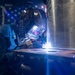 A welder is actively welding at the base of a large cylindrical metal structure, producing a bright arc flash. The worker is wearing a decorative auto-darkening welding helmet branded 'ARCCAPTAIN' and a blue welding jacket. An American flag is visible in the background, suggesting a workshop or fabrication shop setting.