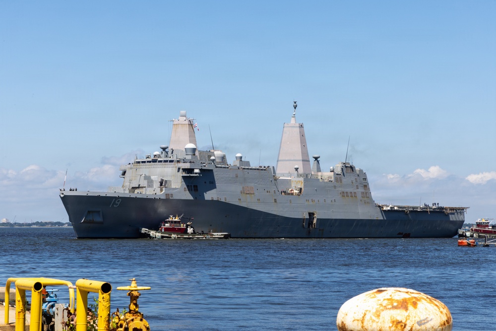 A large US Navy amphibious transport dock ship bearing hull number 19 is visible on open water, assisted by a red tugboat. The vessel features the distinctive angular superstructure and twin mast configuration characteristic of San Antonio-class ships. The image is taken from a dock or pier area with yellow industrial piping and a rusted mooring bollard visible in the foreground.