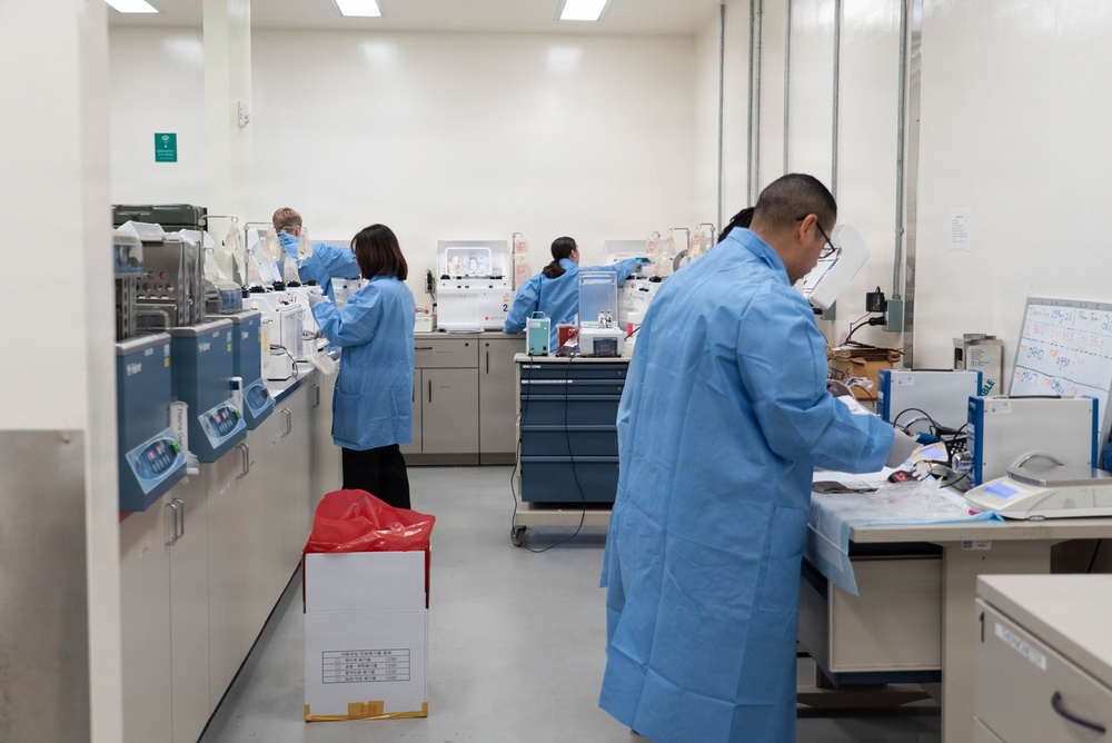 A clinical or research laboratory setting showing multiple technicians in blue lab coats working at various workstations. The lab is well-equipped with analytical instruments, centrifuges, and other laboratory equipment arranged along benches. At least four personnel are visible, each engaged in different laboratory tasks.