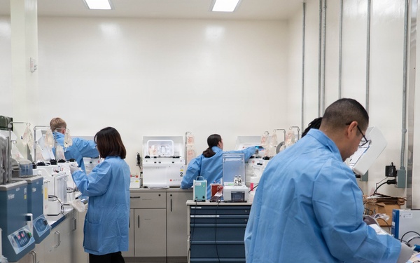 A clinical or research laboratory setting showing multiple technicians in blue lab coats working at various workstations. The lab is well-equipped with analytical instruments, centrifuges, and other laboratory equipment arranged along benches. At least four personnel are visible, each engaged in different laboratory tasks.