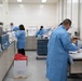 A clinical or research laboratory setting showing multiple technicians in blue lab coats working at various workstations. The lab is well-equipped with analytical instruments, centrifuges, and other laboratory equipment arranged along benches. At least four personnel are visible, each engaged in different laboratory tasks.